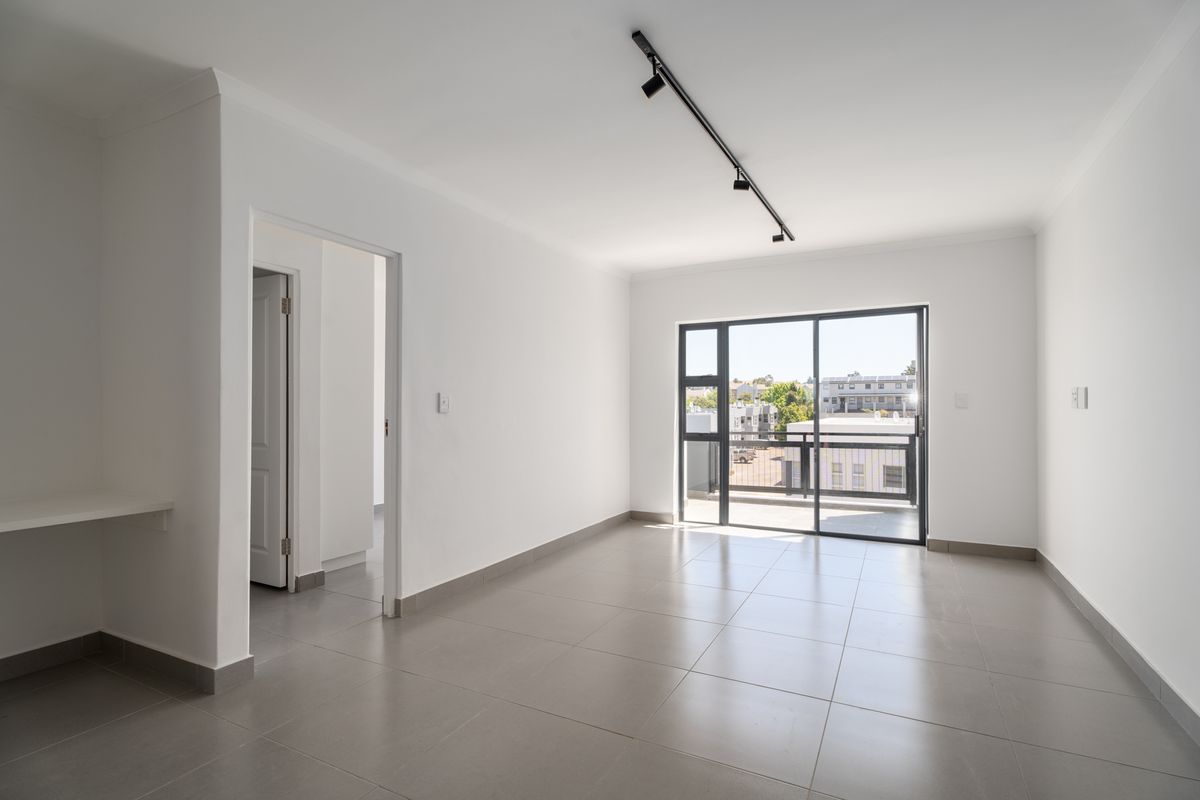 A bright, empty living area featuring neutral tiled flooring, modern track lighting, and a sliding glass door leading to a balcony.