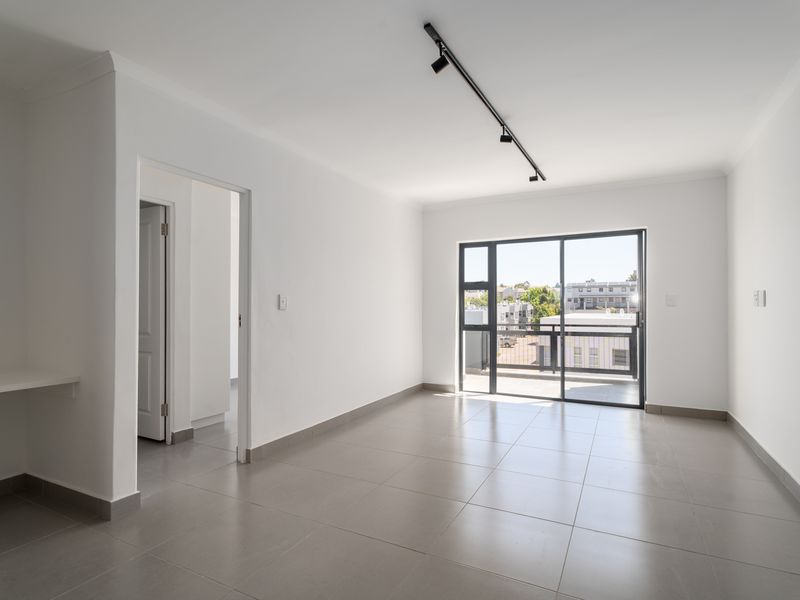 A bright, empty living area featuring neutral tiled flooring, modern track lighting, and a sliding glass door leading to a balcony.