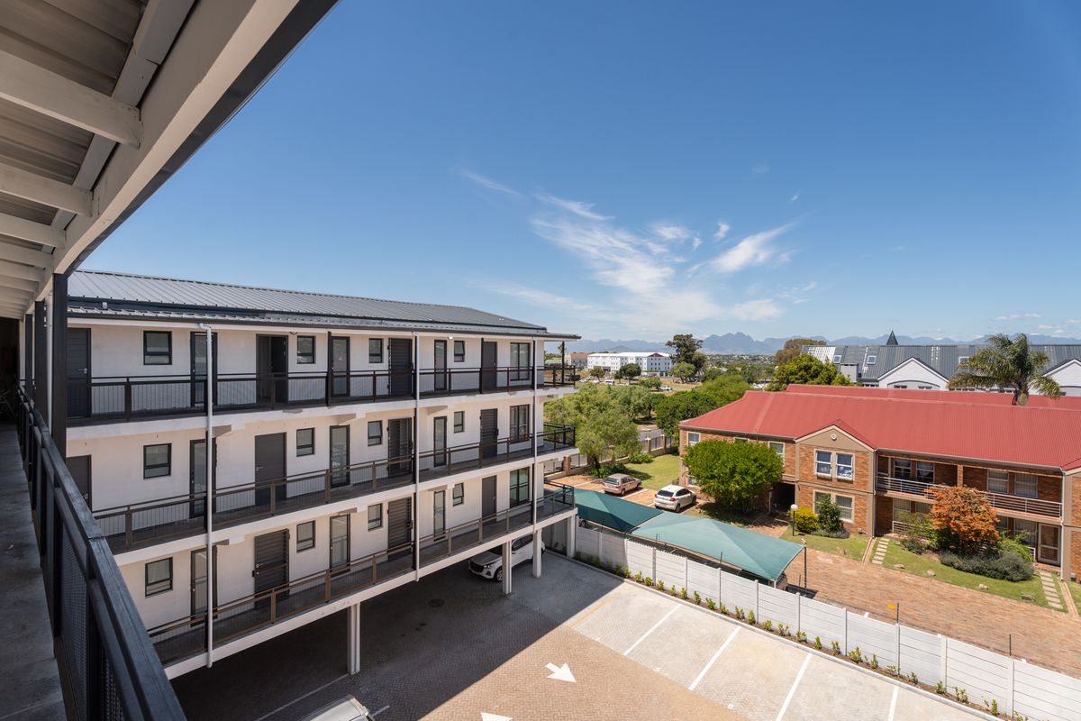 An elevated view of the apartment complex exterior, featuring multi-level walkways, balcony railings, and a paved parking area with surrounding landscaping.