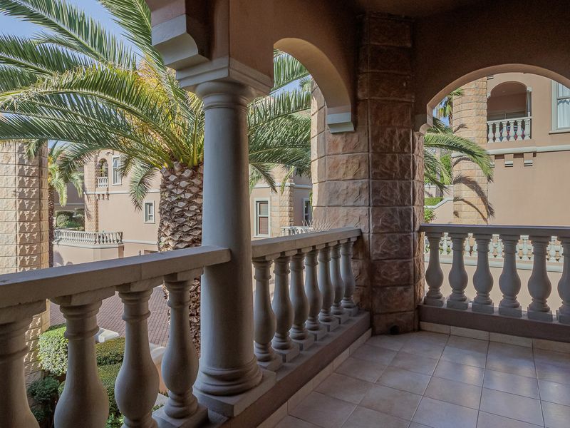 A covered balcony featuring stone-clad columns, arched architectural details, and a classic balustrade, overlooking the apartment complex grounds.