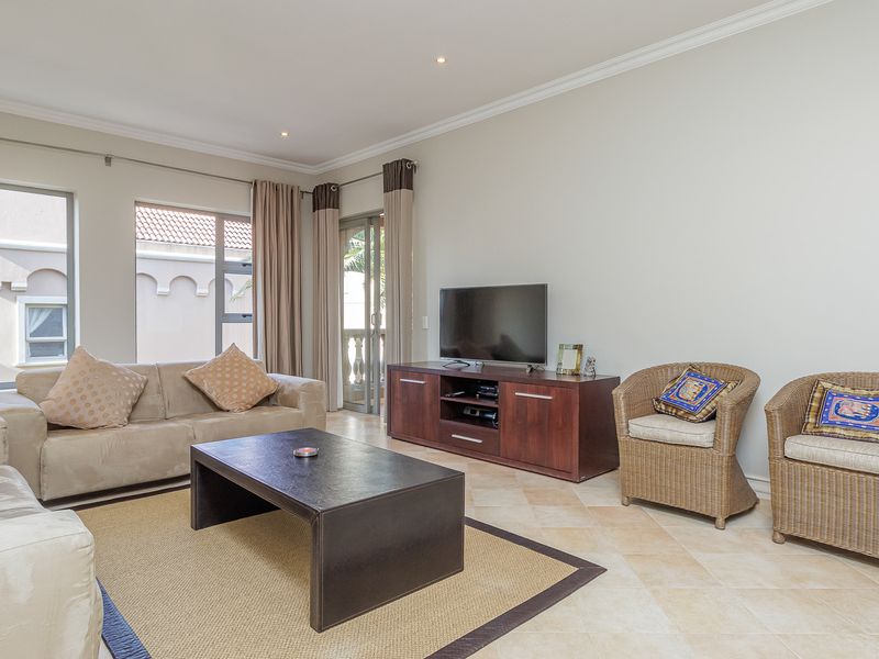 A bright and spacious living room featuring neutral tiled flooring, recessed downlighting, and large windows providing natural light and access to the balcony.