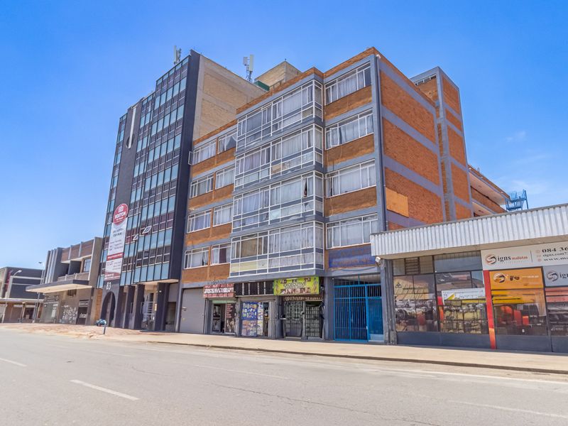 Street-level view of a multi-story apartment building featuring a brick facade, enclosed balconies, and ground-floor commercial retail spaces.