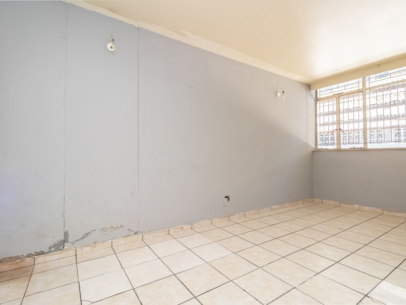 An empty living area featuring tiled flooring and a window with security bars, requiring cosmetic attention to the walls and finishes.