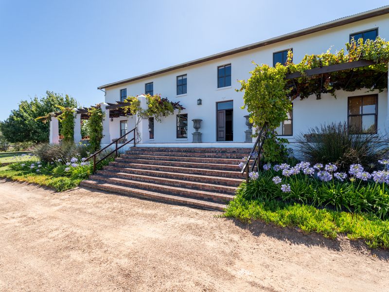 A charming farmhouse exterior featuring a wide brick staircase leading to the main entrance, complemented by a covered pergola and established garden landscaping.
