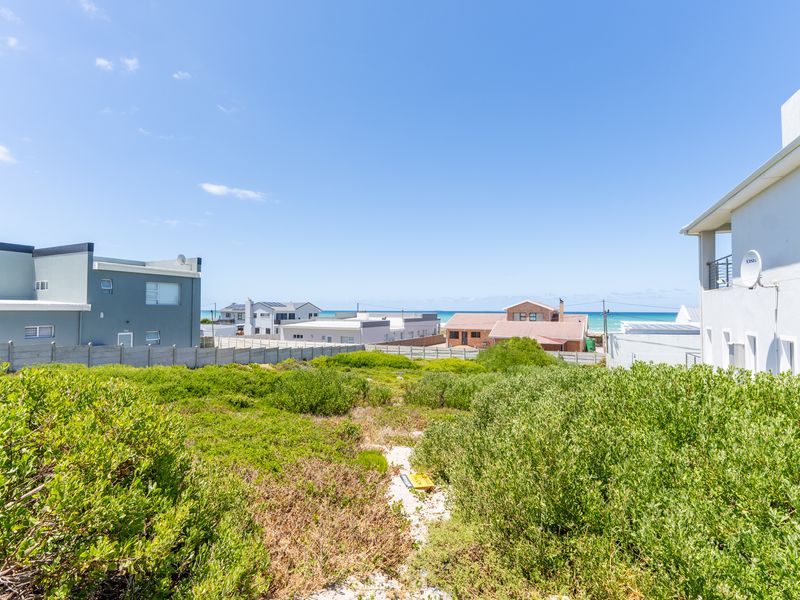 A vacant residential plot featuring coastal vegetation with surrounding residential properties and a glimpse of the ocean in the distance.
