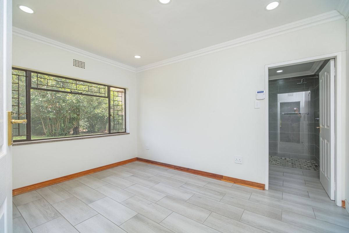 A bright, empty bedroom featuring modern wood-look tile flooring, crown molding, recessed lighting, and an en-suite bathroom with a glass-enclosed shower.