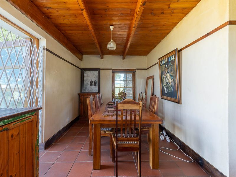 Dining room featuring a rustic exposed timber beam ceiling, terracotta tiled flooring, and large windows with security gates, offering a traditional farm-style aesthetic.
