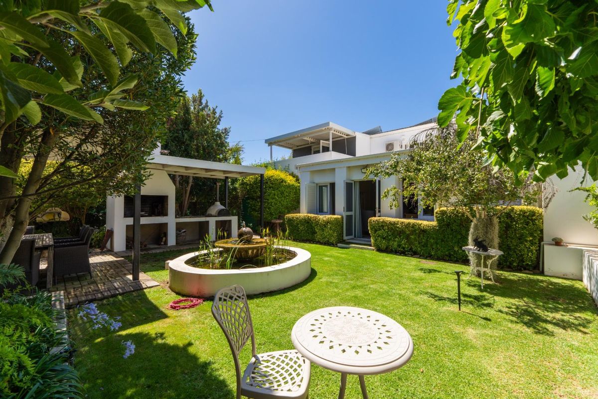 A well-maintained rear garden featuring a paved patio with a built-in braai, a central water feature, and established landscaping.