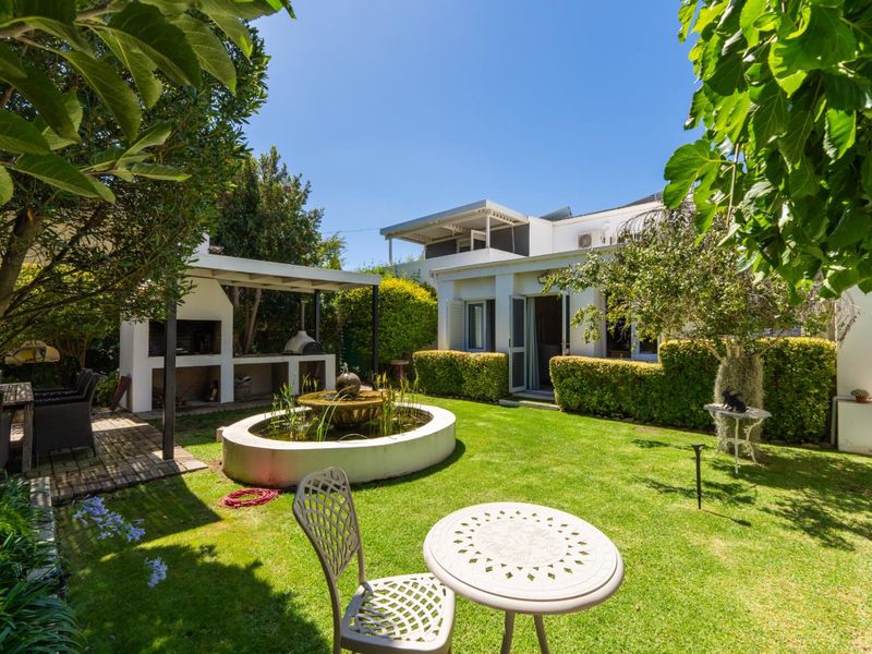 A well-maintained rear garden featuring a paved patio with a built-in braai, a central water feature, and established landscaping.