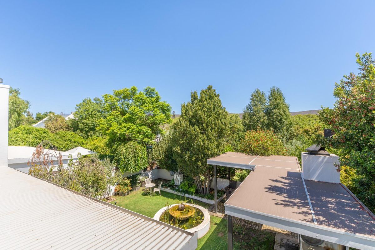 Aerial view of a well-maintained garden featuring a central water feature, covered patio area, and mature trees, viewed from an elevated position.