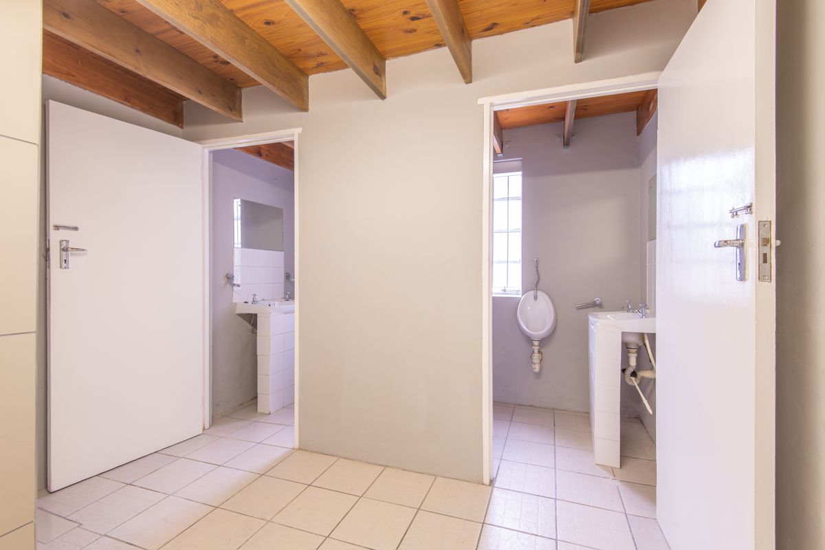 A functional office restroom area featuring two separate stalls with pedestal sinks and a wall-mounted urinal, finished with tiled flooring and exposed timber beams.