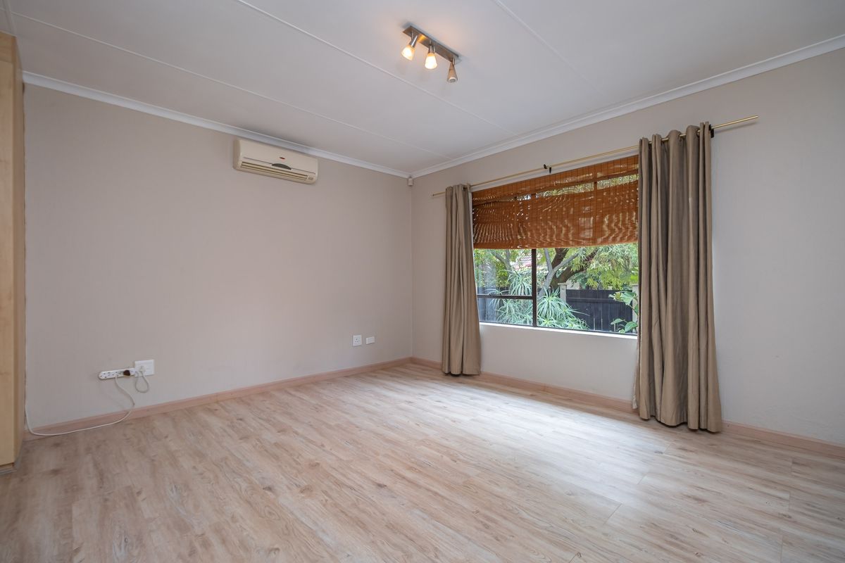 A bright, empty bedroom featuring light wood-look laminate flooring, a wall-mounted air conditioning unit, and large windows with neutral curtains.