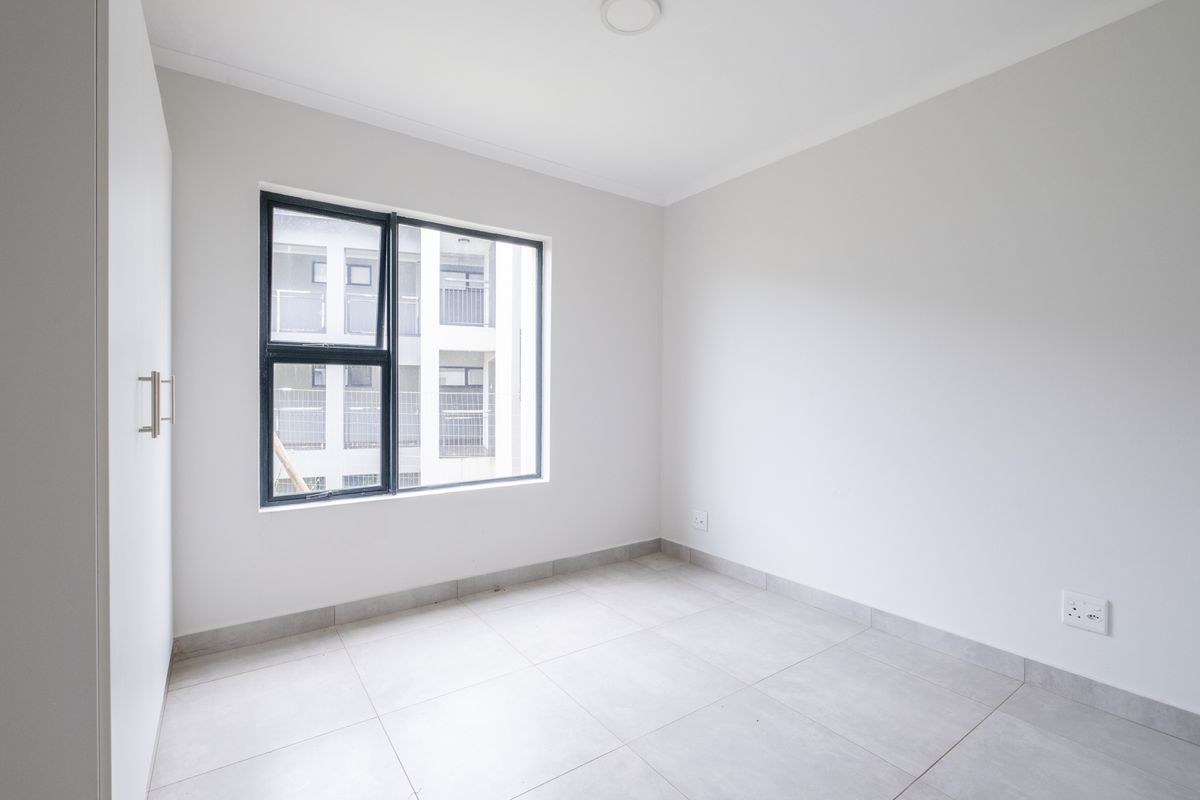 A bright, empty bedroom featuring neutral tiled flooring, a built-in wardrobe, and a large window with a modern black frame.
