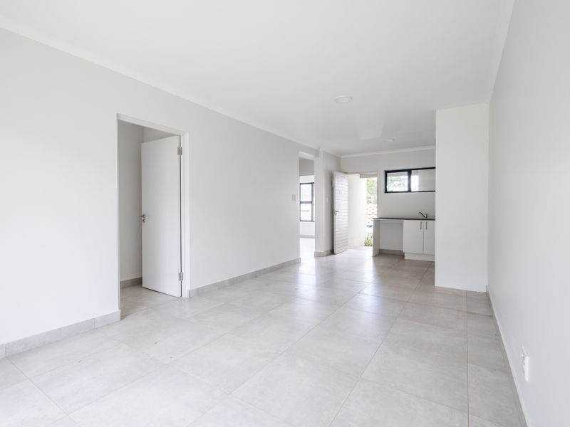 A bright, open-plan living area featuring neutral floor tiles, clean white walls, and a compact kitchen corner with built-in cabinetry and a sink.