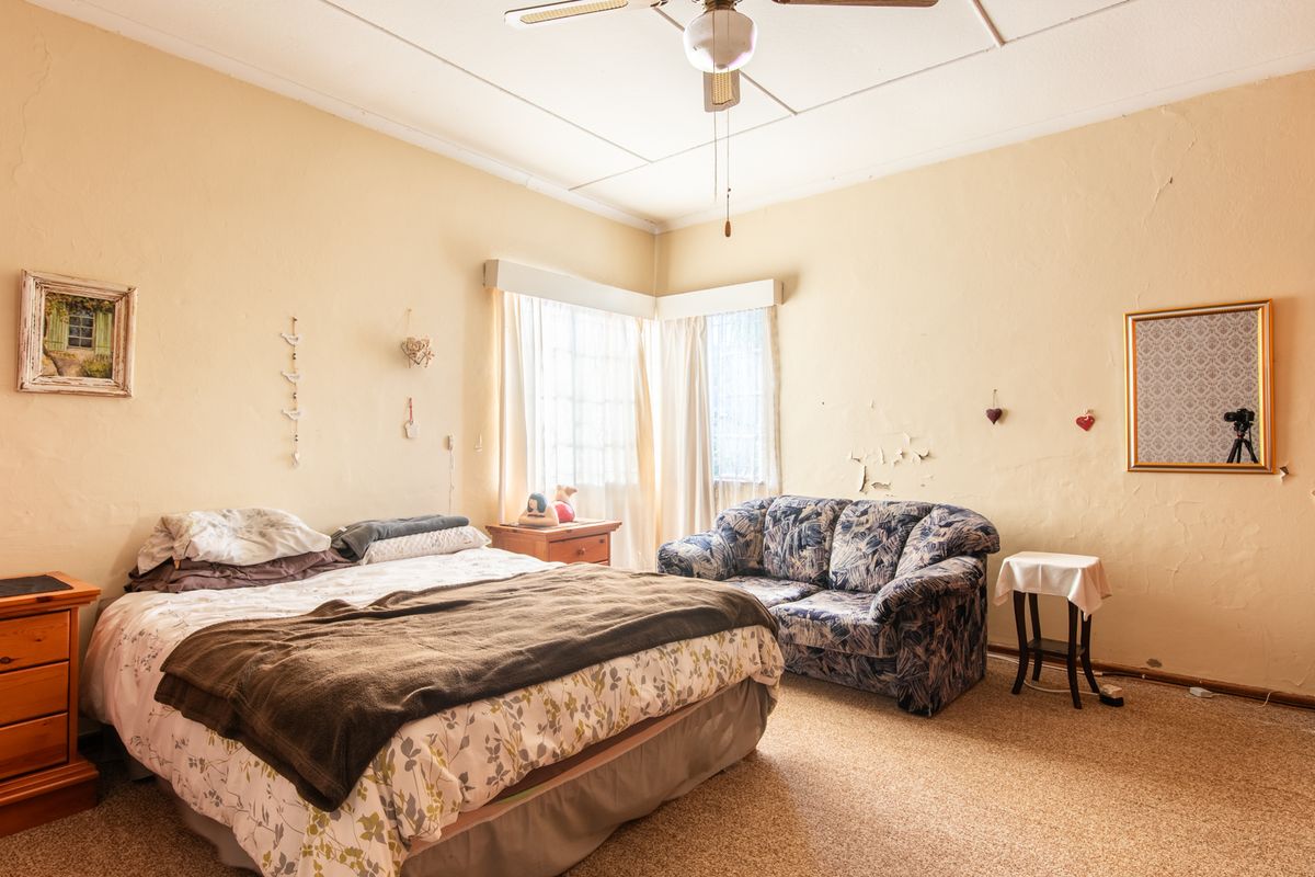 A spacious bedroom featuring built-in wooden cabinetry, a ceiling fan, and large windows, currently requiring cosmetic updates to the wall finishes and flooring.