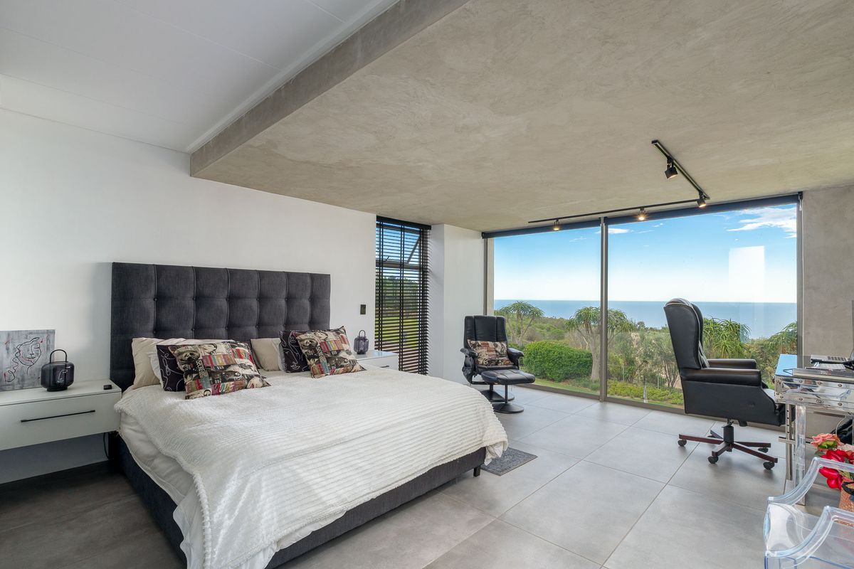 Modern bedroom featuring floor-to-ceiling glass sliding doors with expansive ocean and coastal views, concrete ceiling detail, and large format floor tiles.