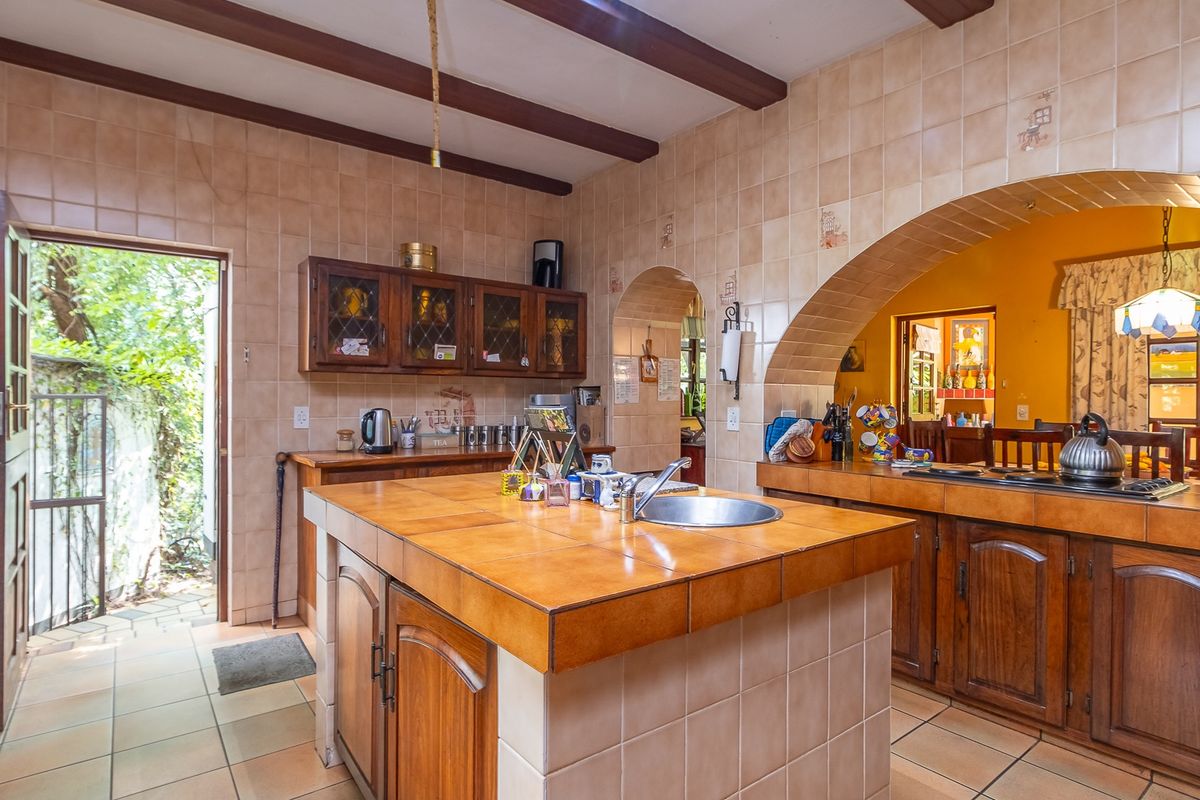 A rustic farm-style kitchen featuring a central island with a built-in sink, tiled countertops, wooden cabinetry, and exposed ceiling beams.