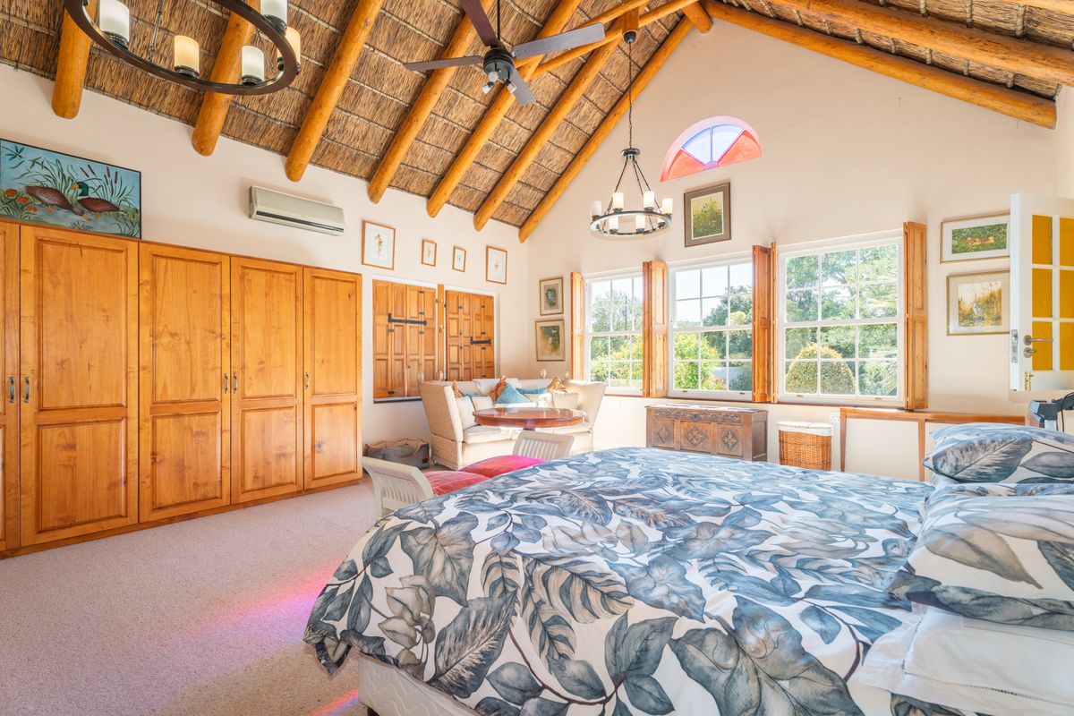 Spacious bedroom featuring high vaulted ceilings with exposed timber rafters, built-in wooden cabinetry, and large windows with timber shutters overlooking the garden.