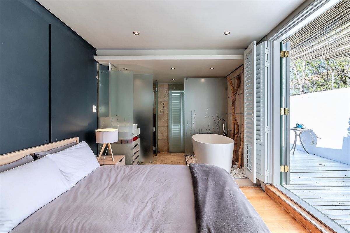 A modern bedroom featuring an open-plan en-suite bathroom with a freestanding bathtub, glass partitions, and direct access to a private wooden deck.