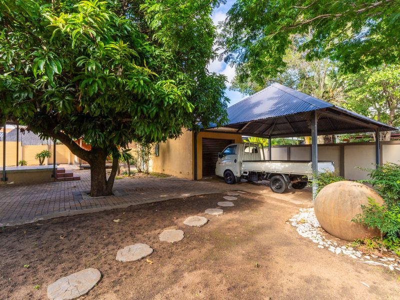 A spacious outdoor area featuring a covered carport, paved pathways, mature trees, and perimeter walling, providing functional exterior space for the property.