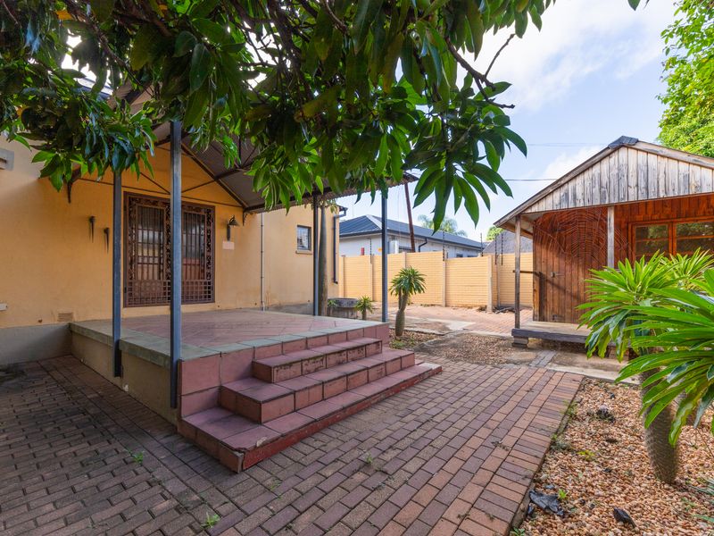 A rear exterior view featuring a covered patio with tiled steps, brick paving, and a separate wooden outbuilding in a landscaped garden setting.