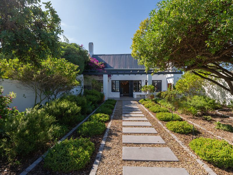 A beautifully landscaped rear garden featuring a stone paver pathway leading to a charming home with a pergola and corrugated metal roof.