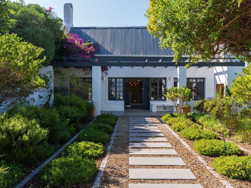 A well-maintained rear exterior view featuring a white facade, corrugated metal roof, pergola, and a stone paver walkway leading to the entrance.