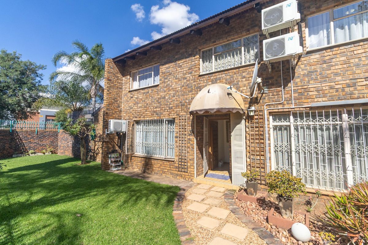 Rear view of a face-brick townhouse featuring a paved walkway, landscaped garden, security gates, and wall-mounted air conditioning units.