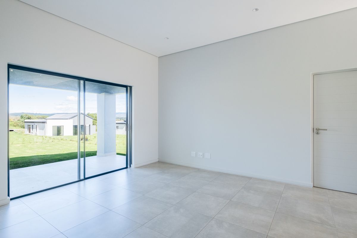A bright, modern, and empty living room featuring large sliding glass doors that open onto a covered patio with garden views.