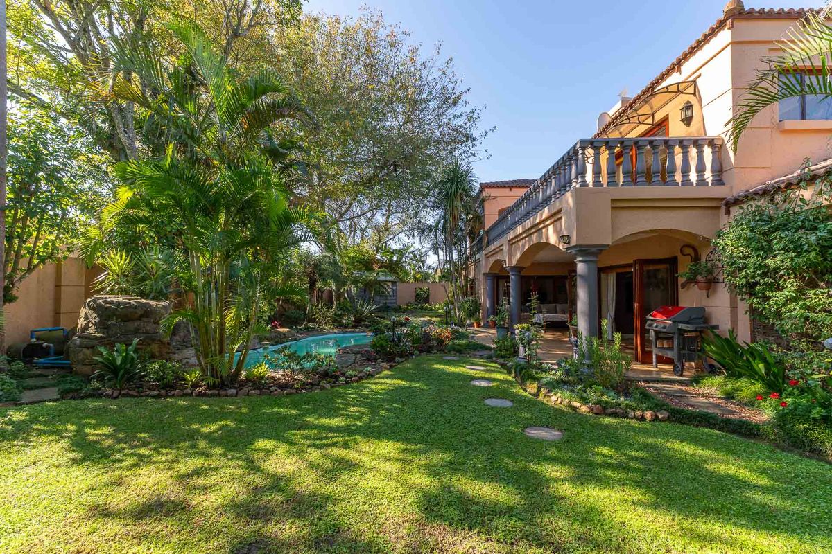 A well-maintained rear garden featuring a swimming pool, lush landscaping, a covered patio area, and an upper-level balcony with a stone balustrade.