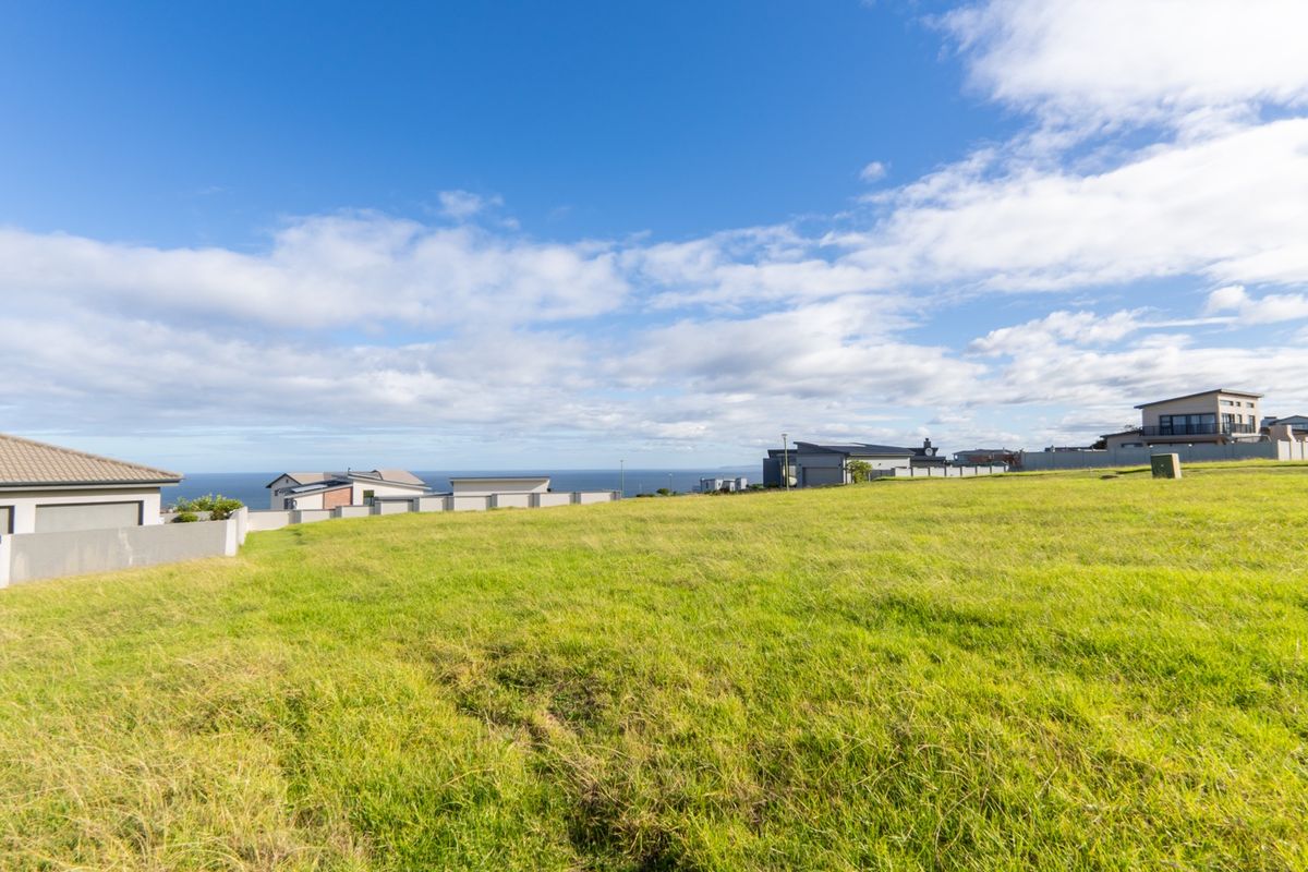 A wide-angle view of a vacant residential plot featuring established grass and boundary walls, with surrounding neighborhood homes and distant ocean views.