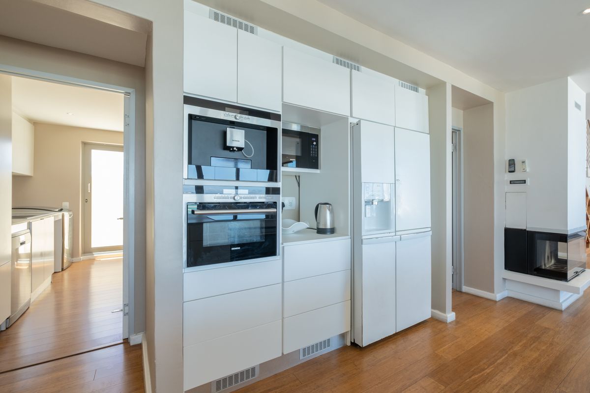 Modern kitchen wall featuring integrated appliances, including a built-in oven, coffee machine, and refrigerator, with sleek white cabinetry and hardwood flooring.