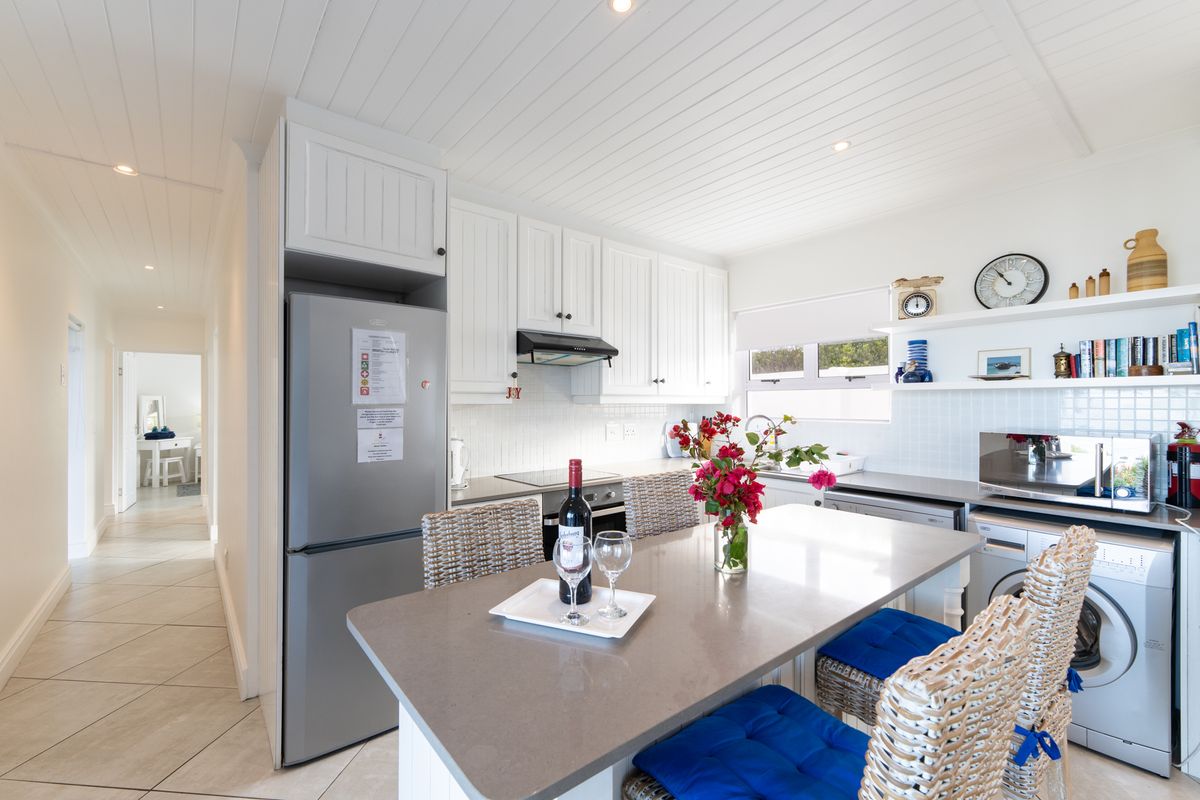 A bright, modern kitchen featuring white cabinetry, stone countertops, a built-in hob, extractor fan, and integrated laundry appliances, with an adjoining breakfast bar.
