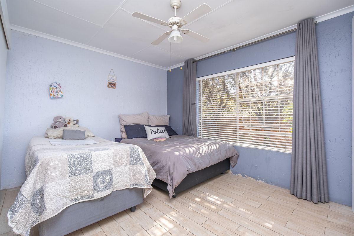 A bedroom featuring tiled flooring, a ceiling fan, and large windows with blinds, providing natural light and a view of the garden.