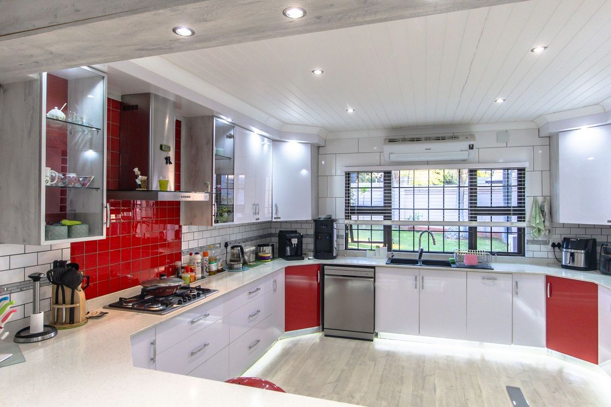Modern kitchen featuring high-gloss cabinetry, a contrasting red tiled backsplash, stone countertops, integrated lighting, and a large window overlooking the garden.