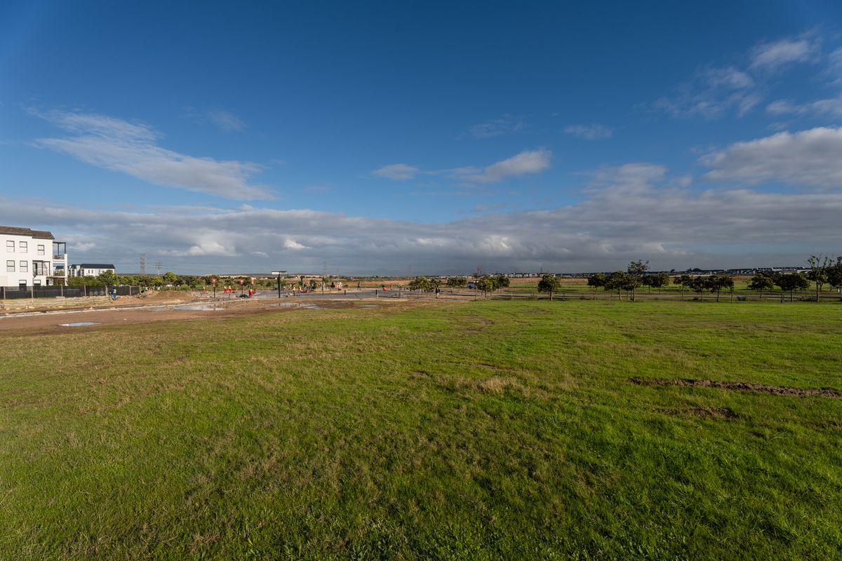 A wide-angle view of a vacant residential plot featuring open grass fields, surrounding neighborhood development, and clear blue skies.
