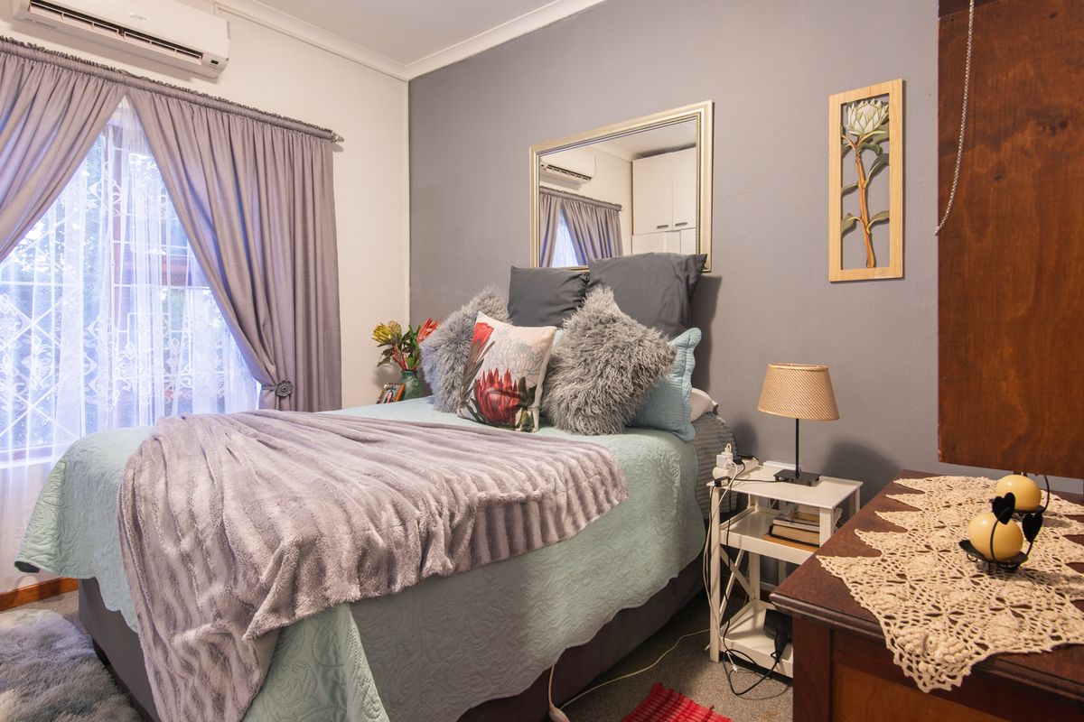 A cozy bedroom featuring a wall-mounted air conditioning unit, large window with curtains, and a neutral grey accent wall.