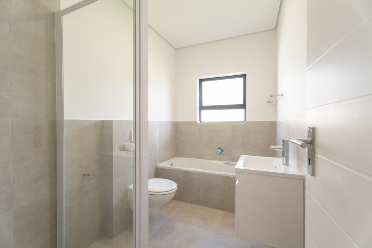 A modern, clean bathroom featuring a built-in bathtub, wall-mounted toilet, vanity basin, and a glass-enclosed shower area with neutral tiling.