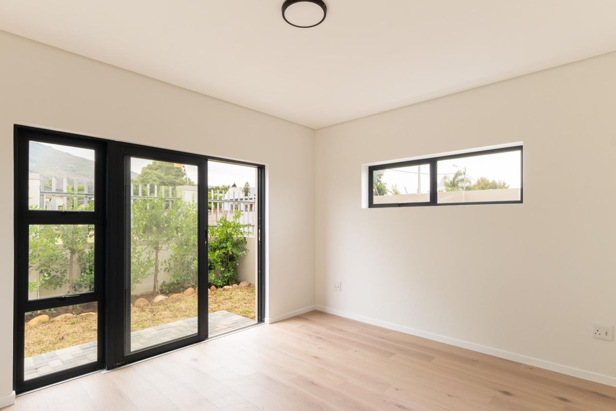 A bright, modern bedroom featuring light wood-look flooring, black-framed sliding glass doors leading to a private garden, and a high-level horizontal window.