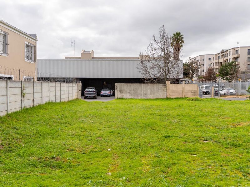 A vacant residential plot featuring a grassed surface, enclosed by concrete boundary walls with security fencing, situated in a developed urban area.