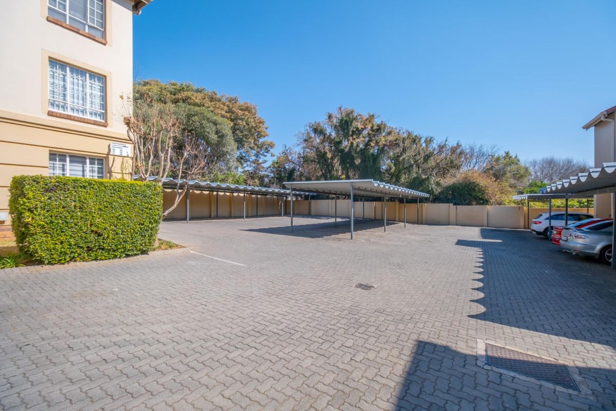 A paved outdoor parking area featuring covered carports, adjacent to the apartment building with established perimeter hedging and boundary walls.