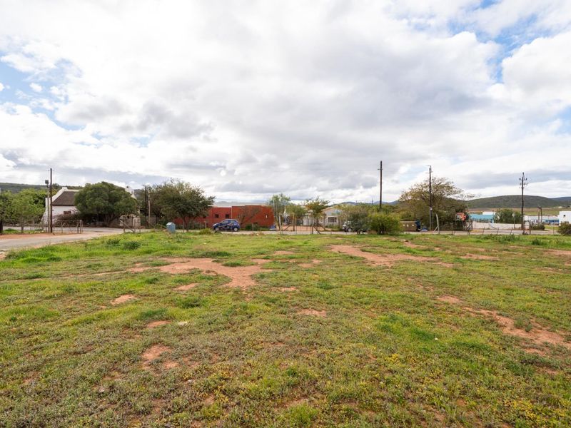 A vacant residential plot featuring open grass and dirt terrain, with surrounding neighborhood infrastructure and utility poles visible in the background.