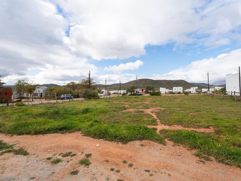 A vacant residential plot of land featuring natural grass and soil, offering a clear view of the surrounding neighborhood and distant mountain landscape.
