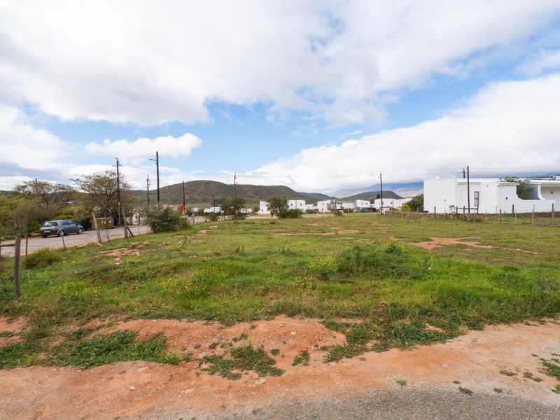 A vacant residential plot of land featuring natural grass coverage, perimeter fencing, and views of the surrounding landscape and nearby residential properties.