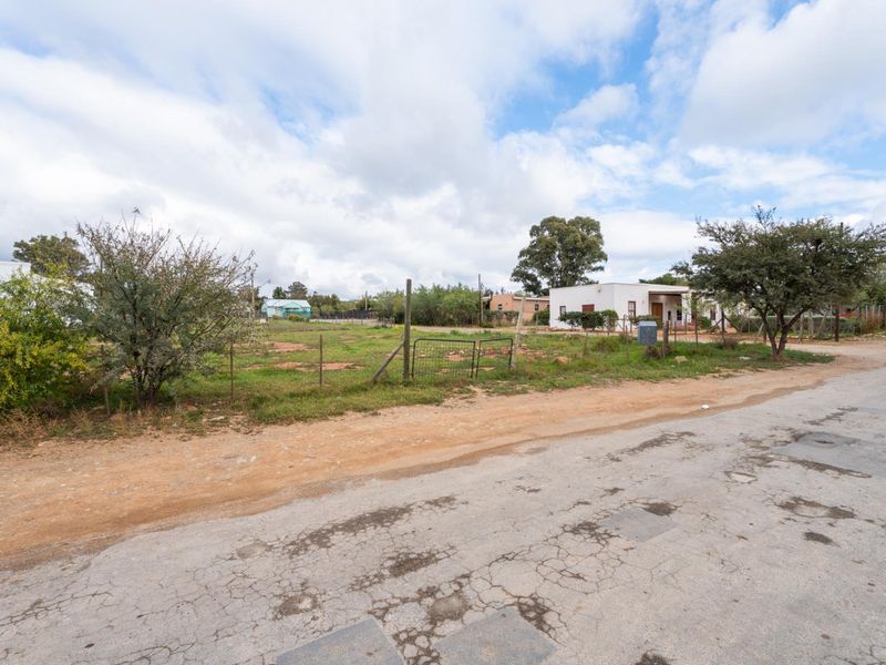 A view of a vacant residential plot from the street, featuring natural vegetation, wire fencing, and surrounding neighborhood context.