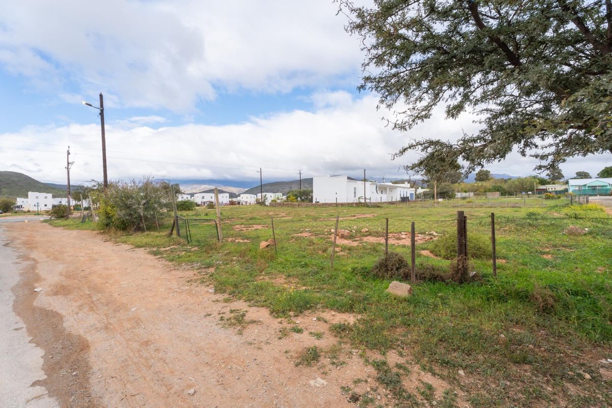 A vacant residential plot featuring natural vegetation, basic wire fencing, and access to a dirt road in a quiet neighborhood setting.