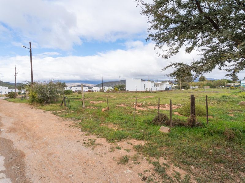 A vacant residential plot featuring natural vegetation, basic wire fencing, and access to a dirt road in a quiet neighborhood setting.