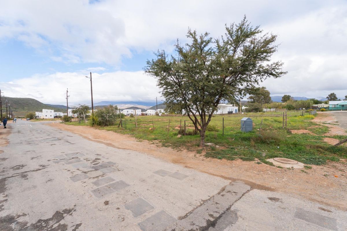 A view of a vacant residential plot from the street, featuring natural vegetation, a perimeter fence, and utility access points.