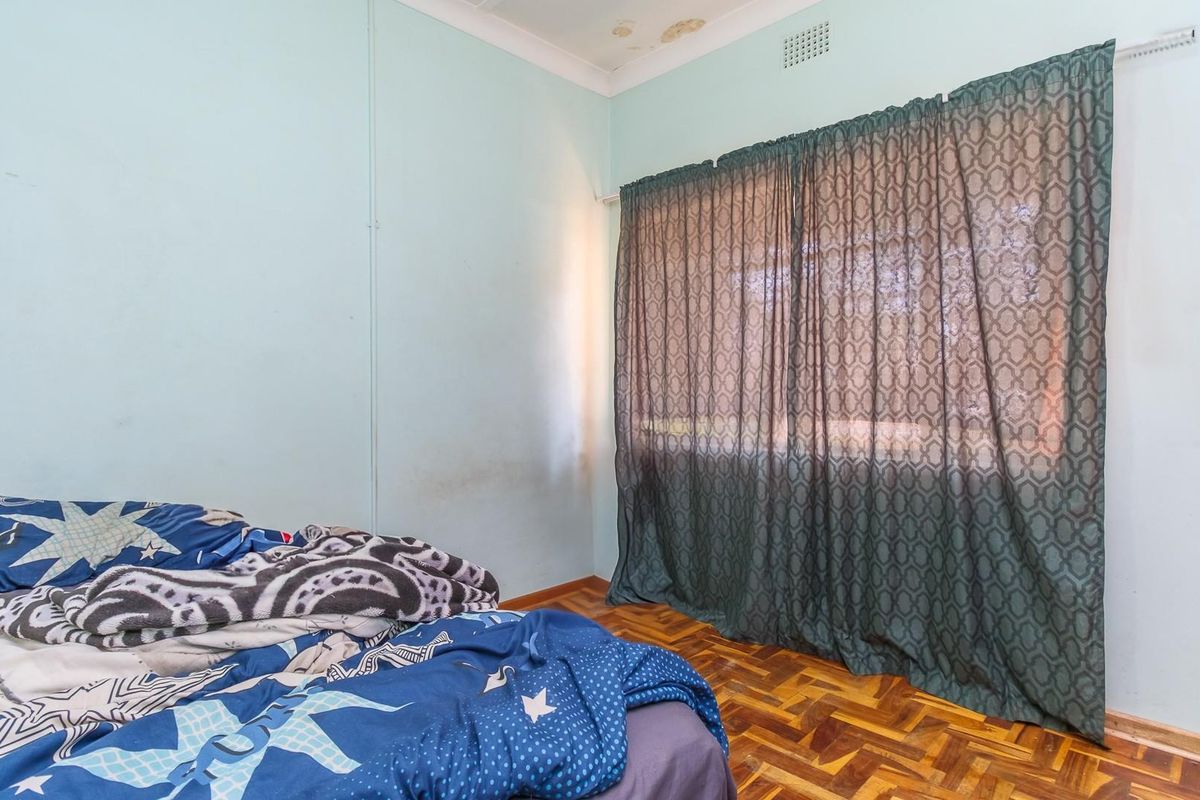 A bedroom featuring parquet flooring, a window with curtain track, and visible signs of ceiling water damage and wall wear.