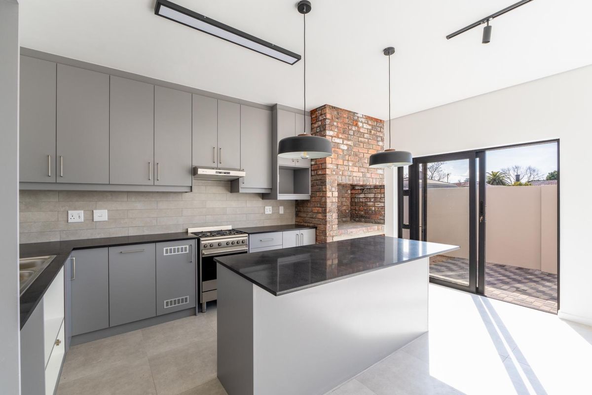 Modern kitchen featuring grey cabinetry, stone countertops, a central island, built-in oven with extractor, and an integrated brick fireplace, opening onto a paved patio.